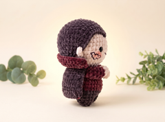 A small crocheted doll wearing a dark purple knit hat and a burgundy knit scarf, standing against a warm cream background with soft green foliage props on both sides.