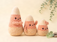 Three crocheted amigurumi candy corn figures in different sizes, styled on a warm beige background with soft green eucalyptus leaves.