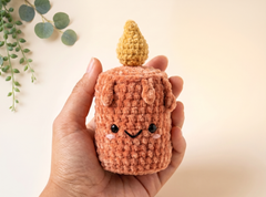 A hand holding a small crocheted plush character shaped like a cute candle, with a peach-pink body, yellow wick, and embroidered face, against a warm cream background with eucalyptus leaves in the corner.