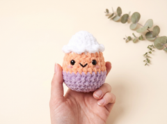A hand holding a small crocheted plush toy shaped like a cute character with a peach face, black embroidered eyes and mouth, a white top, and a lavender bottom, against a warm beige background with eucalyptus leaves in the corner.