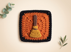 A square crocheted textile piece featuring a stylized broom made from layered yellow, brown, and orange yarn, set against a black border on a warm beige background with decorative green leaves in the corners.