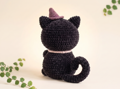 A black crocheted cat plush toy with a purple pointed hat and a light pink collar, shown from the back against a warm cream background with decorative green leaves.