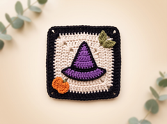 A square handmade crochet coaster featuring a purple witch hat, an orange pumpkin, and a green leaf on a cream background with a black border, styled with eucalyptus leaves on a beige surface.