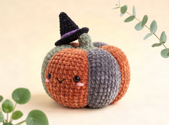 A cute crocheted pumpkin doll with a witch hat, featuring orange, gray, and green sections, set against a warm beige background with decorative eucalyptus leaves.