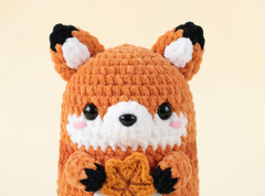A close-up of a crocheted plush fox toy with orange fur, a white face, black ears and paws, and a small yellow-orange leaf in its front paws, set against a soft cream background.