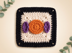 A square crocheted textile piece with a black border, cream inner field, orange circular center, and two purple oval side accents, photographed flat on a light beige background with eucalyptus leaves around the edges.