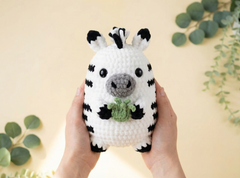 A crocheted zebra plush toy being held in two hands against a warm beige background with green eucalyptus leaves in the corners.