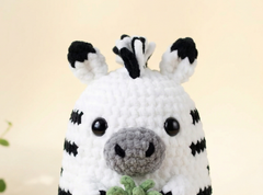 A close-up of a crocheted zebra plush toy with black and white stripes, a gray snout, and black eyes, holding a small green leaf against a soft beige background.
