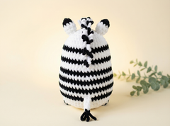 A crocheted zebra-patterned pouch or cover with black and white stripes, shown on a warm cream background with a small green sprig in the corner.