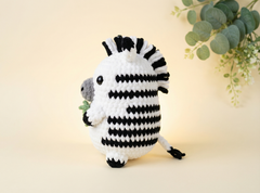 A crocheted zebra plush toy with black and white stripes, a gray snout, and a small green leaf in its front hooves, standing against a warm cream background with eucalyptus leaves in the upper right corner.