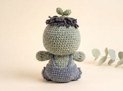 A handmade crocheted plush toy shaped like a baby plant, shown from the back. The toy has a sage green textured body, dark gray yarn hair with two small green sprouts on top, and blue-gray overalls.